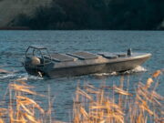 Small aluminum motorboat gliding on a blue lake with a rocky shore in the background, grasses in the foreground.