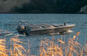 Meet Katran X1.2, Ukraine’s swarming drone carrier Small aluminum motorboat gliding on a blue lake with a rocky shore in the background, grasses in the foreground.