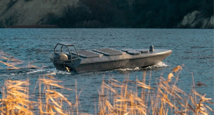 Small aluminum motorboat gliding on a blue lake with a rocky shore in the background, grasses in the foreground.