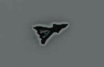 Small black irregular triangular sticker with a white outline on a gray surface, facing to the right.