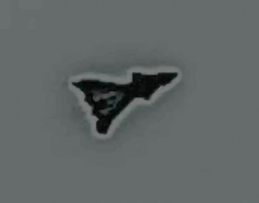 Small black irregular triangular sticker with a white outline on a gray surface, facing to the right.