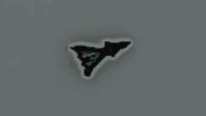 Small black irregular triangular sticker with a white outline on a gray surface, facing to the right.