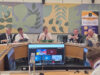 Panel of six men seated around a curved conference table in a formal meeting room, with a Stephen Hoffman Consulting banner in the background and large decorative wall panels behind them.