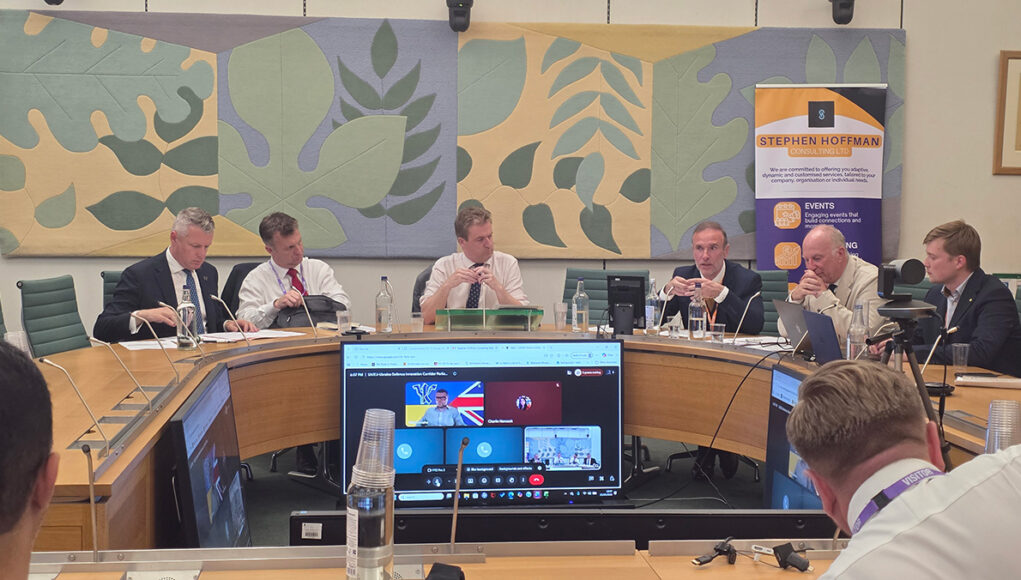 Panel of six men seated around a curved conference table in a formal meeting room, with a Stephen Hoffman Consulting banner in the background and large decorative wall panels behind them.