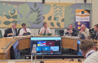 Panel of six men seated around a curved conference table in a formal meeting room, with a Stephen Hoffman Consulting banner in the background and large decorative wall panels behind them.