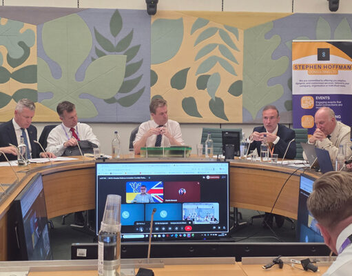 Panel of six men seated around a curved conference table in a formal meeting room, with a Stephen Hoffman Consulting banner in the background and large decorative wall panels behind them.
