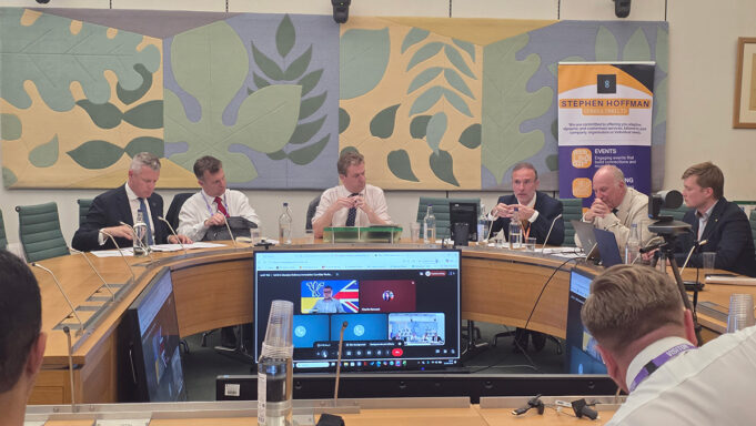 Panel of six men seated around a curved conference table in a formal meeting room, with a Stephen Hoffman Consulting banner in the background and large decorative wall panels behind them.