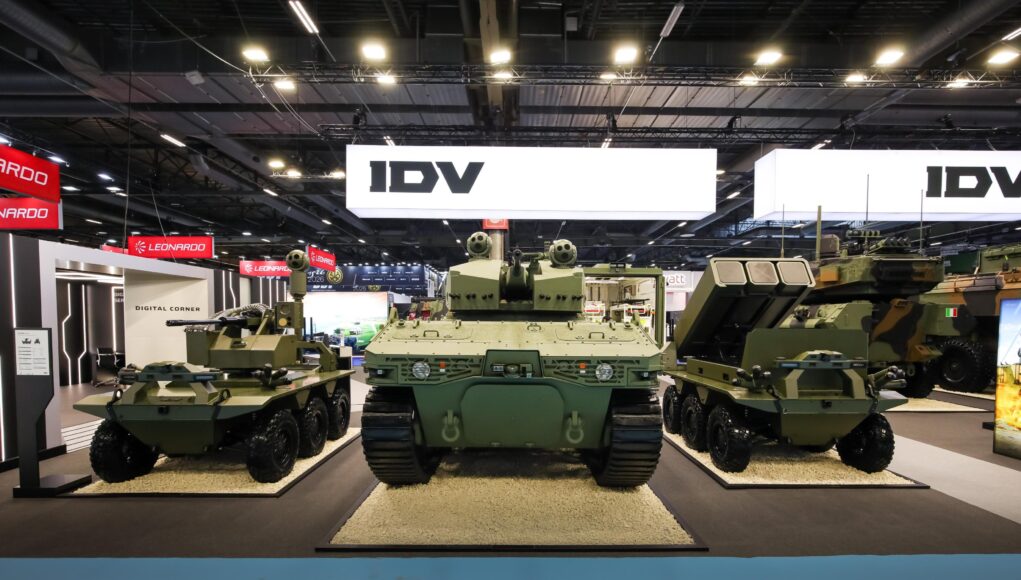 pro-COSmzSOz Military tanks on display at an exhibition hall with bright overhead lighting and large IDV signs in the background.