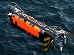 Orange and gray underwater robotic device suspended above dark blue water, with a metal frame and yellow/black fins visible on the side.