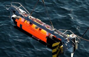 Support deal planned for Royal Navy mine disposal system Orange and gray underwater robotic device suspended above dark blue water, with a metal frame and yellow/black fins visible on the side.