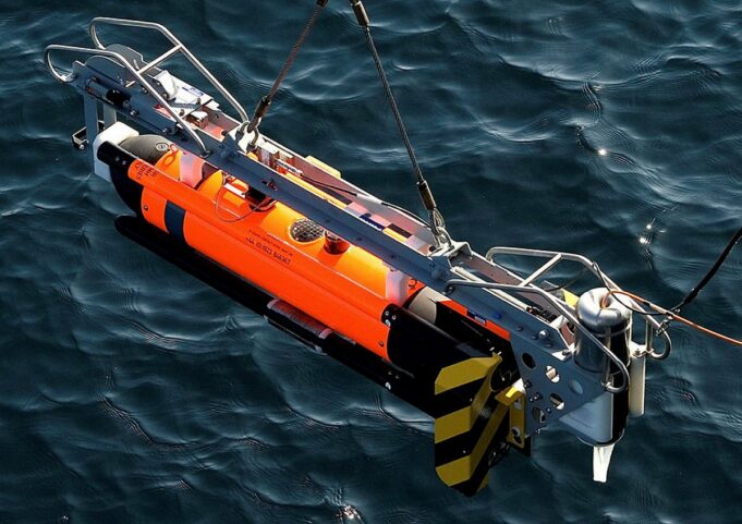 Orange and gray underwater robotic device suspended above dark blue water, with a metal frame and yellow/black fins visible on the side.