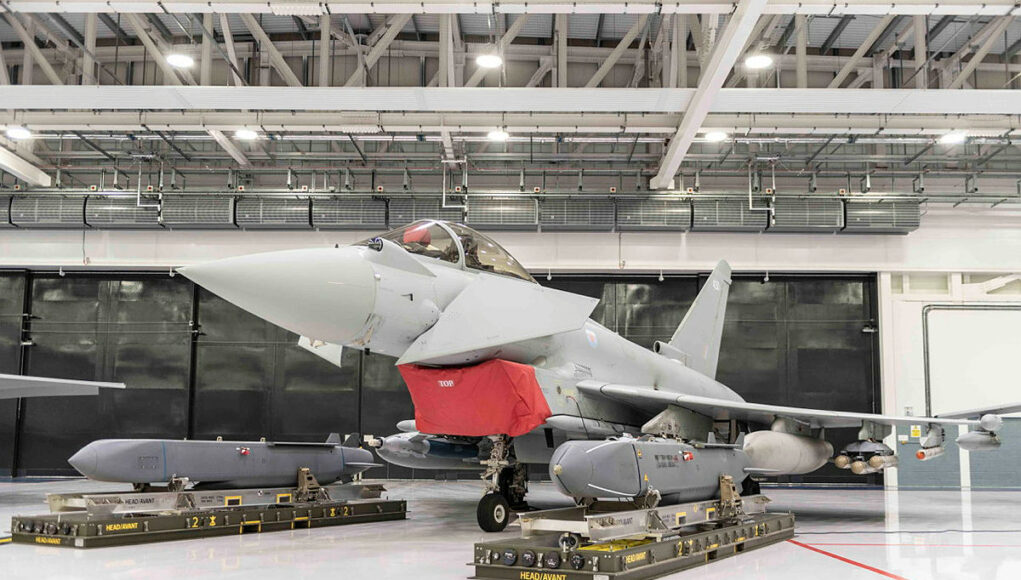 Gray military fighter jet in a hangar on transport pallets, with a red protective cover over the intake and maintenance equipment nearby.