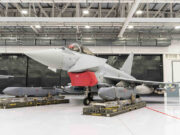 Gray military fighter jet in a hangar on transport pallets, with a red protective cover over the intake and maintenance equipment nearby.