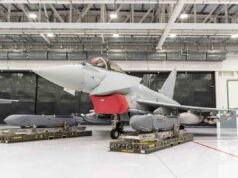 Gray military fighter jet in a hangar on transport pallets, with a red protective cover over the intake and maintenance equipment nearby.
