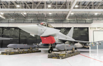 Gray military fighter jet in a hangar on transport pallets, with a red protective cover over the intake and maintenance equipment nearby.