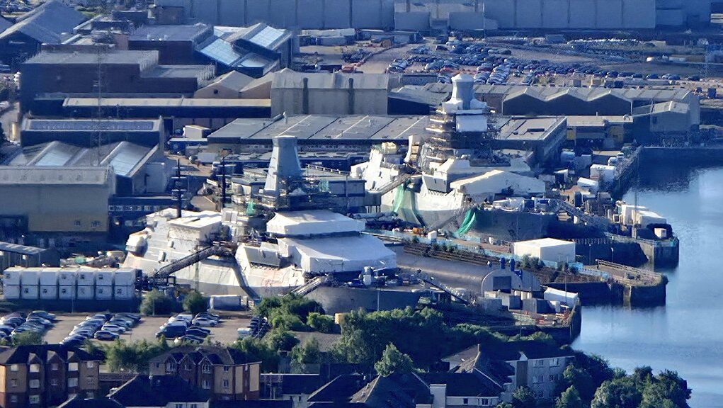 A waterfront industrial site with numerous white tents and temporary structures near the harbor, surrounded by warehouses and parked cars.