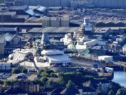 A waterfront industrial site with numerous white tents and temporary structures near the harbor, surrounded by warehouses and parked cars.