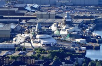 A waterfront industrial site with numerous white tents and temporary structures near the harbor, surrounded by warehouses and parked cars.