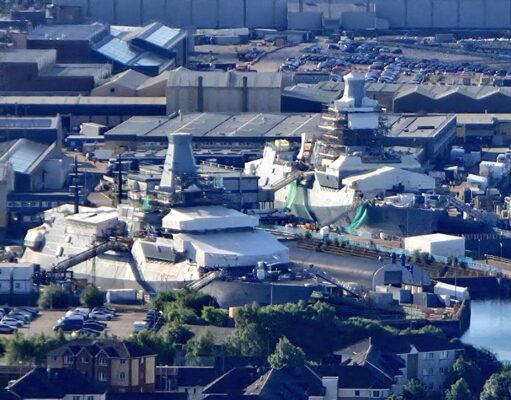 A waterfront industrial site with numerous white tents and temporary structures near the harbor, surrounded by warehouses and parked cars.