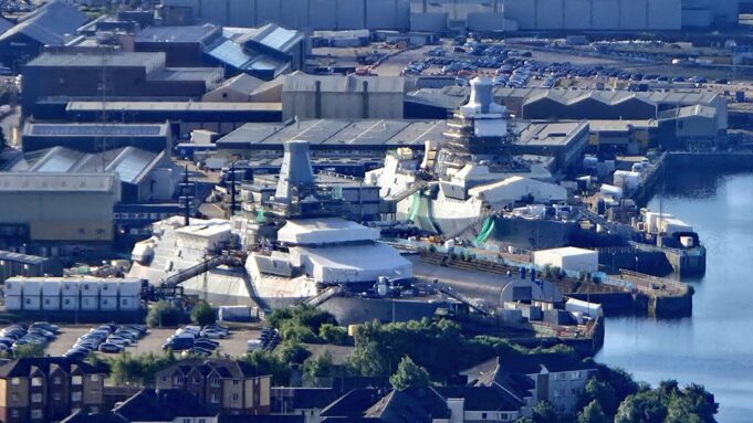 A waterfront industrial site with numerous white tents and temporary structures near the harbor, surrounded by warehouses and parked cars.