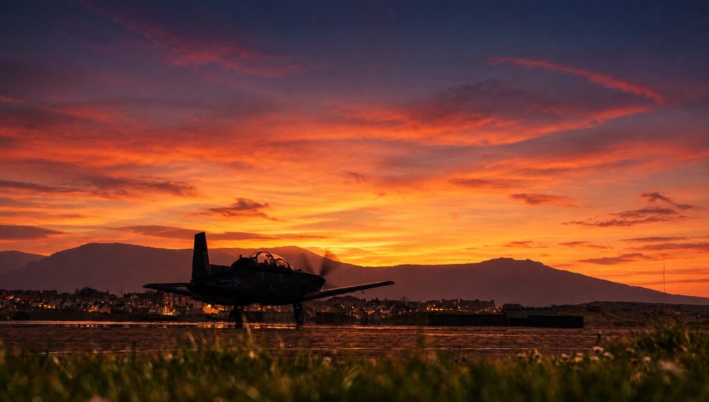 Silhouette of a small aircraft on a runway at sunset, with an orange sky and distant mountains.