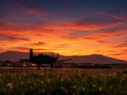 Silhouette of a small aircraft on a runway at sunset, with an orange sky and distant mountains.