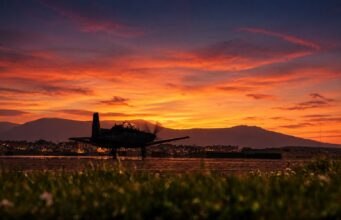 Silhouette of a small aircraft on a runway at sunset, with an orange sky and distant mountains.
