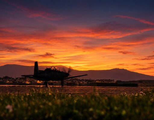Silhouette of a small aircraft on a runway at sunset, with an orange sky and distant mountains.