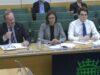 Three panelists seated at a conference table with nameplates for Andrew Kinniburgh, Lucia Retter, and Max Warner in front of them.