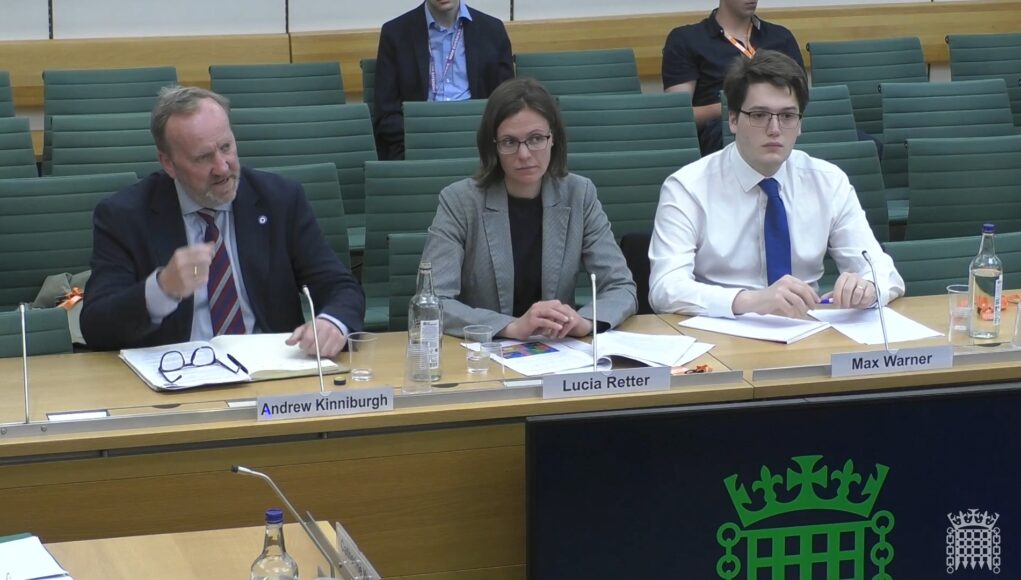Three panelists seated at a conference table with nameplates for Andrew Kinniburgh, Lucia Retter, and Max Warner in front of them.
