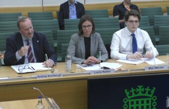 Three panelists seated at a conference table with nameplates for Andrew Kinniburgh, Lucia Retter, and Max Warner in front of them.