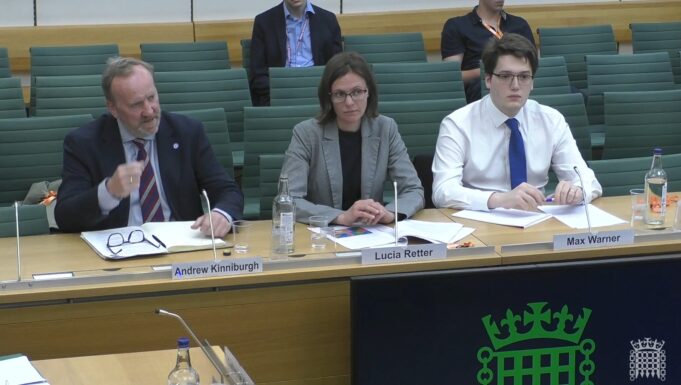 Three panelists seated at a conference table with nameplates for Andrew Kinniburgh, Lucia Retter, and Max Warner in front of them.