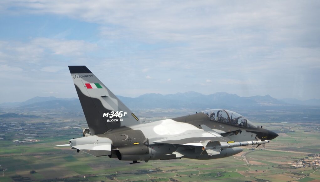 Military jet in flight, Italian M-346F trainer with flag on tail against a rural valley and mountains far below.