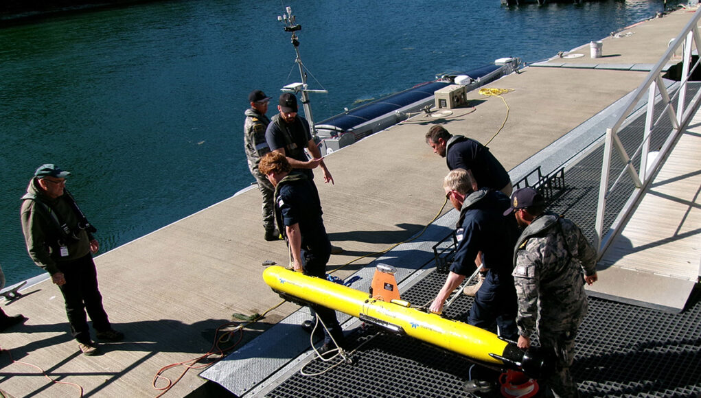 Crew members in work gear secure a bright yellow torpedo-shaped device on a dock by the water, with cables and equipment around it.
