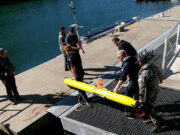 Royal Navy tests seabed defence tech with AUKUS allies Crew members in work gear secure a bright yellow torpedo-shaped device on a dock by the water, with cables and equipment around it.