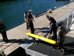 Royal Navy tests seabed defence tech with AUKUS allies Crew members in work gear secure a bright yellow torpedo-shaped device on a dock by the water, with cables and equipment around it.