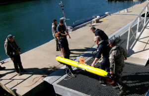 Royal Navy tests seabed defence tech with AUKUS allies Crew members in work gear secure a bright yellow torpedo-shaped device on a dock by the water, with cables and equipment around it.