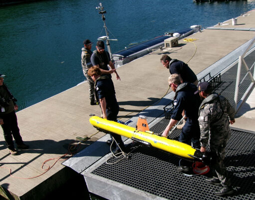Crew members in work gear secure a bright yellow torpedo-shaped device on a dock by the water, with cables and equipment around it.
