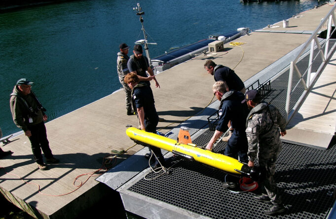 Royal Navy tests seabed defence tech with AUKUS allies Crew members in work gear secure a bright yellow torpedo-shaped device on a dock by the water, with cables and equipment around it.
