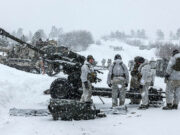 BAE lands £4M UK deal for artillery and gun components Soldiers in white winter gear stand around a large artillery cannon in a snowy battlefield, preparing or inspecting the weapon as snow falls heavily.