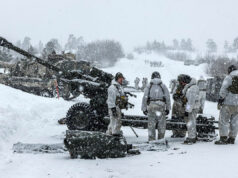 BAE lands £4M UK deal for artillery and gun components Soldiers in white winter gear stand around a large artillery cannon in a snowy battlefield, preparing or inspecting the weapon as snow falls heavily.