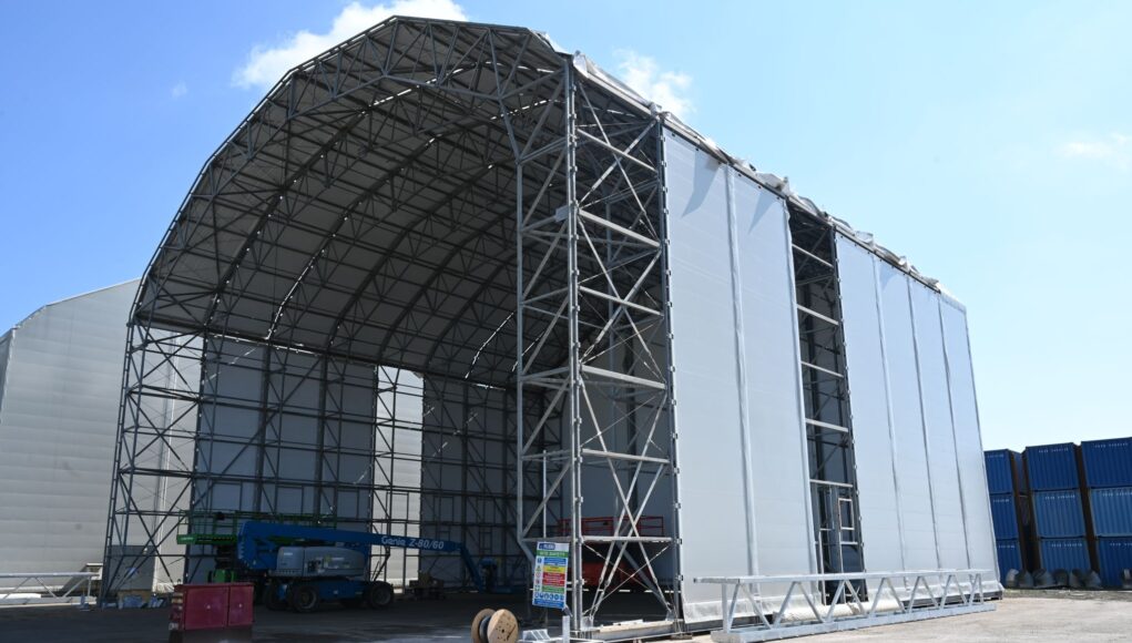 1784880328212 Construction site: a large steel-framed warehouse under a curved roof with white fabric walls against a blue sky.