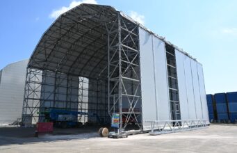 Construction site: a large steel-framed warehouse under a curved roof with white fabric walls against a blue sky.