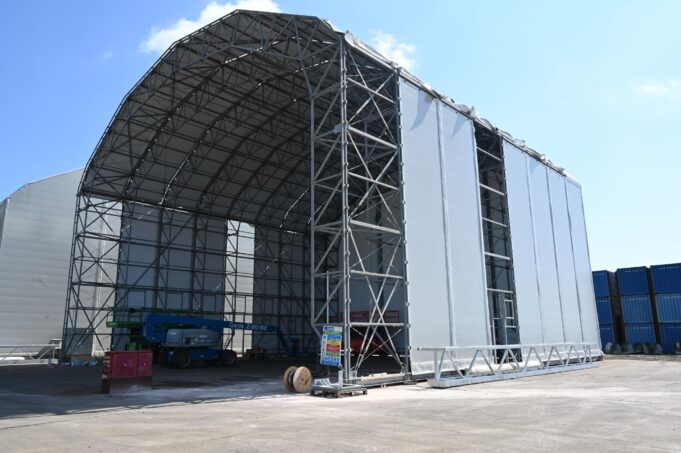 Construction site: a large steel-framed warehouse under a curved roof with white fabric walls against a blue sky.