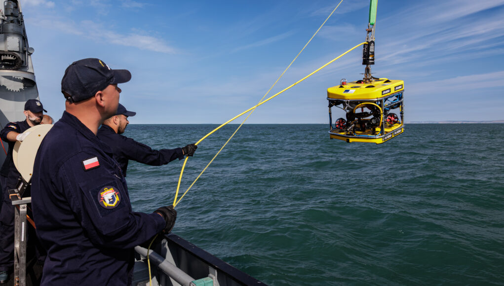 Polish naval personnel lower a yellow underwater drone (ROV) into the sea from a ship, secured by yellow cables against a blue sky and water.