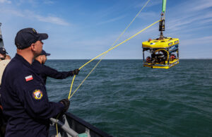 Polish naval personnel lower a yellow underwater drone (ROV) into the sea from a ship, secured by yellow cables against a blue sky and water.
