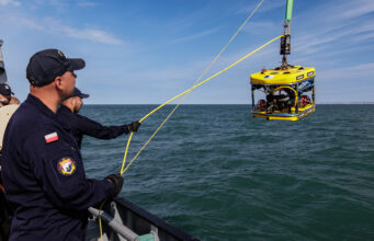 Sea Breeze brings allied EOD divers to Portland waters Polish naval personnel lower a yellow underwater drone (ROV) into the sea from a ship, secured by yellow cables against a blue sky and water.