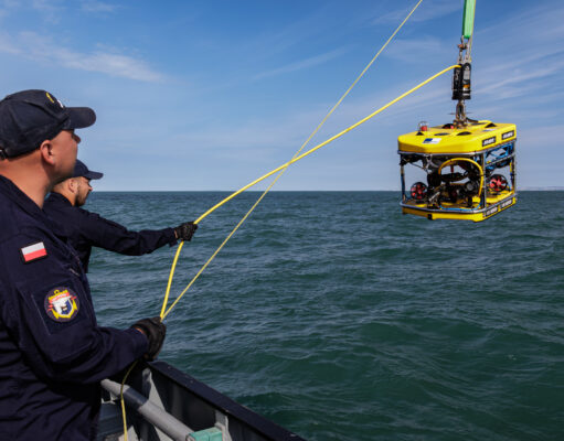 Sea Breeze brings allied EOD divers to Portland waters Polish naval personnel lower a yellow underwater drone (ROV) into the sea from a ship, secured by yellow cables against a blue sky and water.