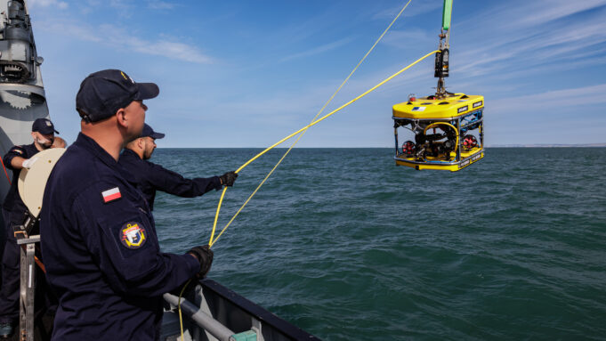 Sea Breeze brings allied EOD divers to Portland waters Polish naval personnel lower a yellow underwater drone (ROV) into the sea from a ship, secured by yellow cables against a blue sky and water.