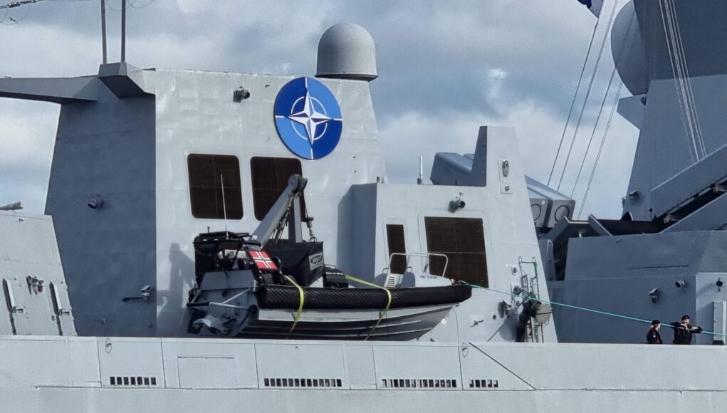 20221013_135431 Gray warship deck with a large blue NATO emblem on the superstructure, lifeboat in front, and a few crew members at the lower right.